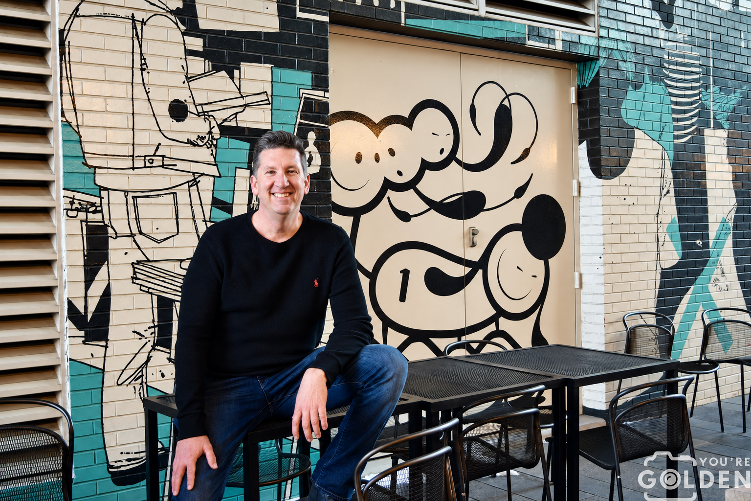 Bradley Cowdrey sitting outside beside a mural, smiling